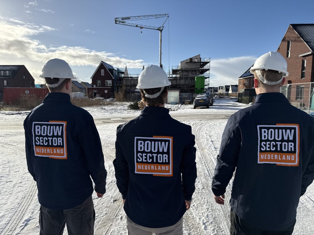 Drie vakmensen van Bouwsector Nederland met witte helm en werkjas met logo lopen over een besneeuwde bouwplaats met nieuwe huizen en een bouwkraan